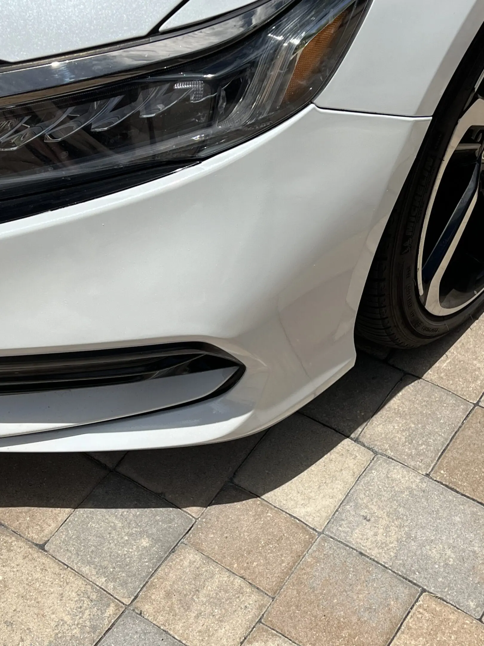 Close-up of a white car's front bumper and wheel on a paved surface.