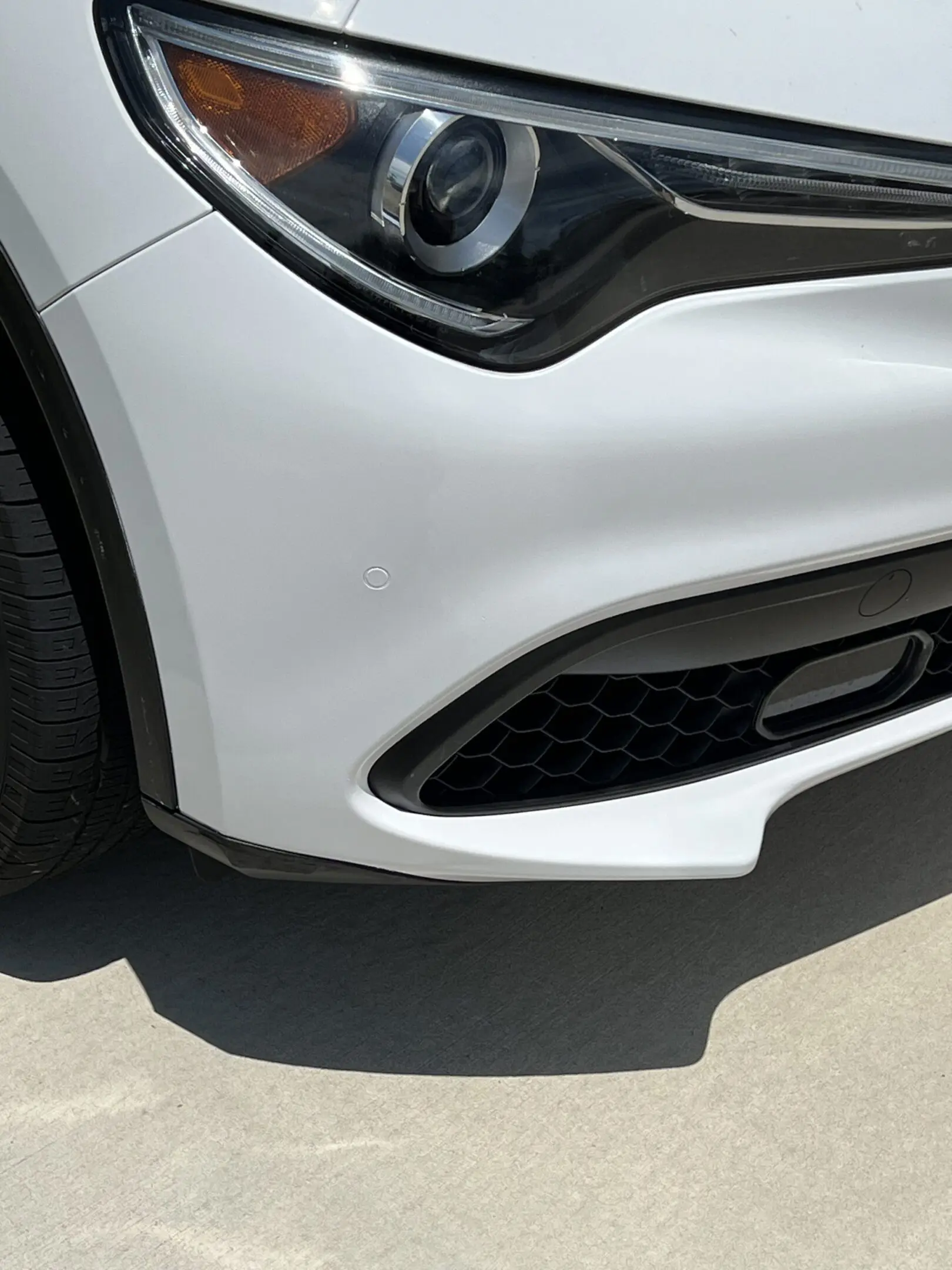 Close-up of a white car's front bumper and headlight under sunlight.